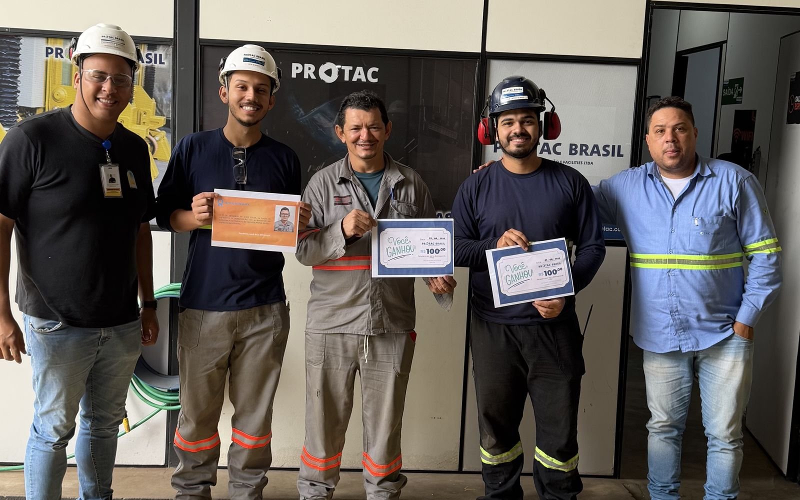 Equipe da PROTAC Brasil com certificados, diante do painel da empresa